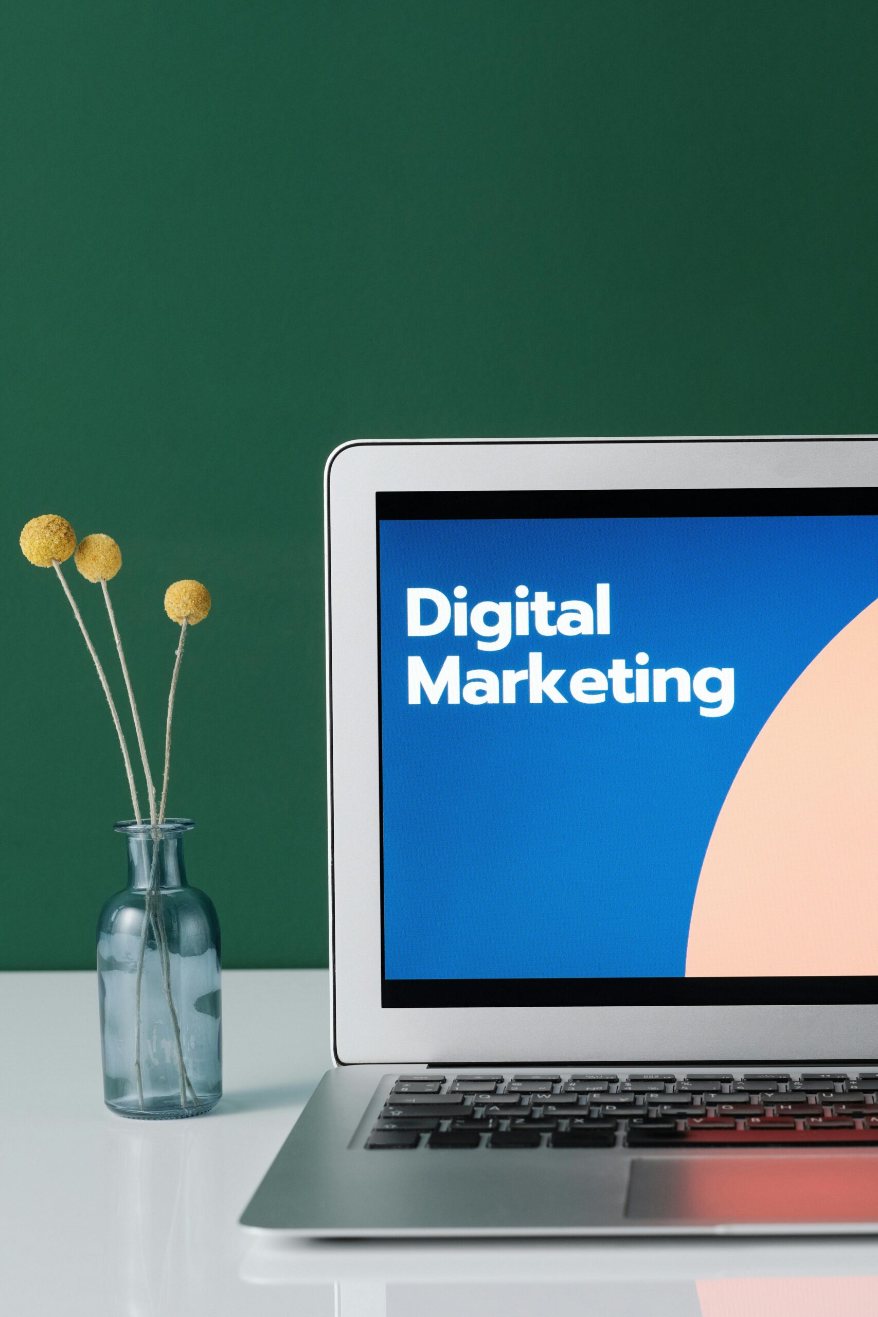 The image shows a minimalist and professional setup. On the right side, there is an open laptop with the screen displaying the text "Digital Marketing" in bold white font over a modern, colorful background with blue and peach tones.