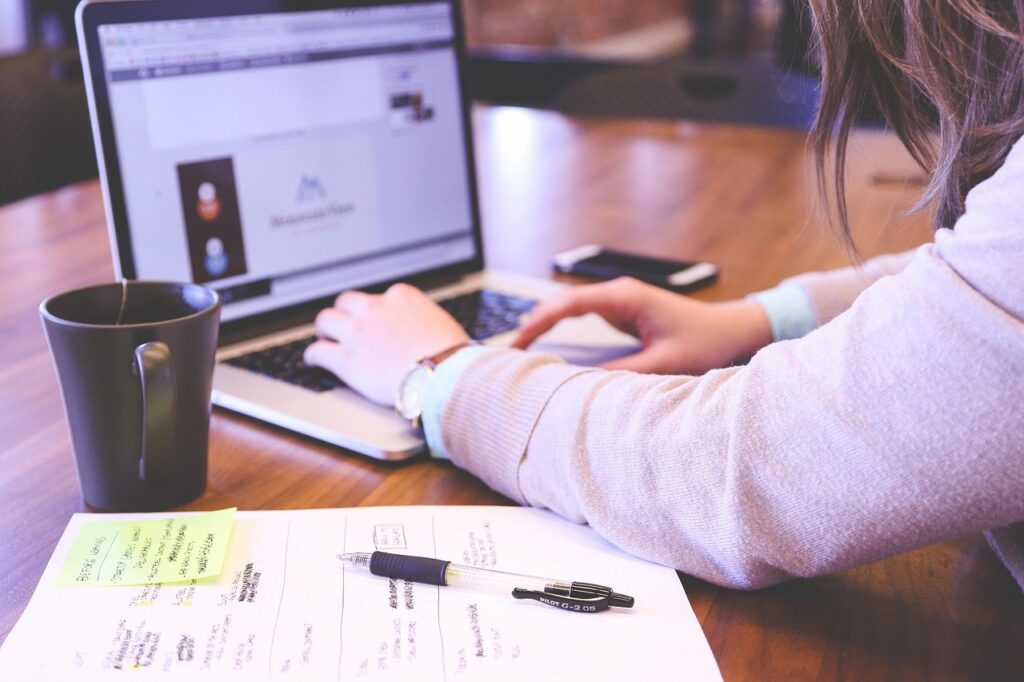The image shows a side view of a woman working at a wooden desk. She is using a laptop, and her hands are on the keyboard, actively typing. On the laptop screen, there appears to be a webpage open, possibly related to design or branding