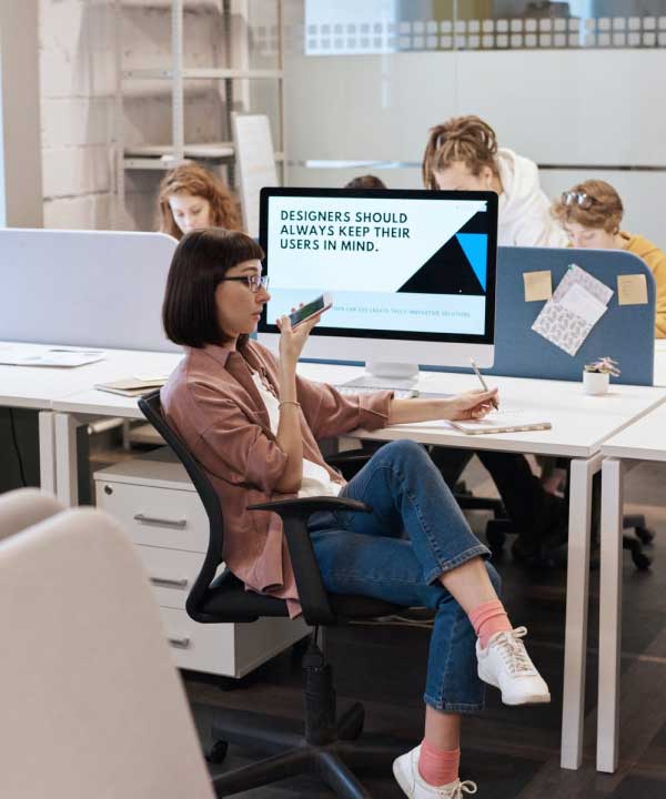 The image shows a modern office workspace with a young woman sitting in a swivel chair, working thoughtfully.