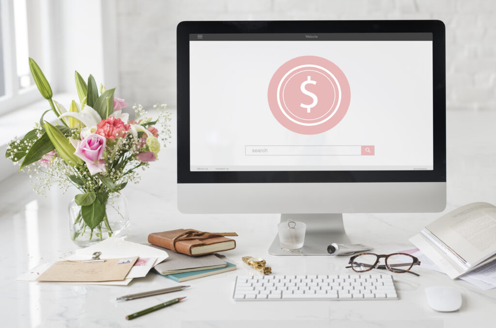 The image showcases a clean and aesthetically pleasing office workspace. At the center is a large desktop computer screen displaying a pink dollar coin icon, symbolizing online finance or possibly a pay-per-click (PPC) advertisement theme.