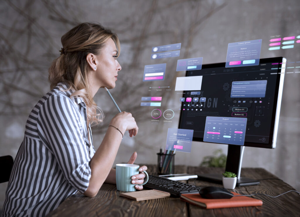 The image shows a woman working at a wooden desk, focused on a computer screen displaying a futuristic user interface design